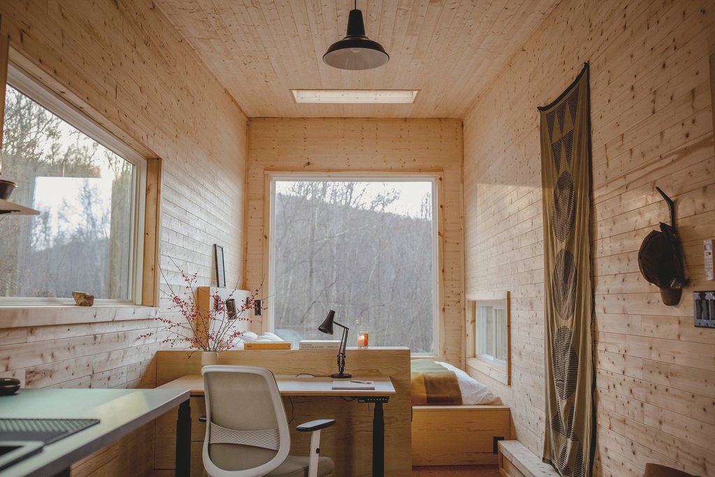 The interior looking down its length, a desk and chair set up to work at under a skylight, with the bed and a window into bare woods beyond